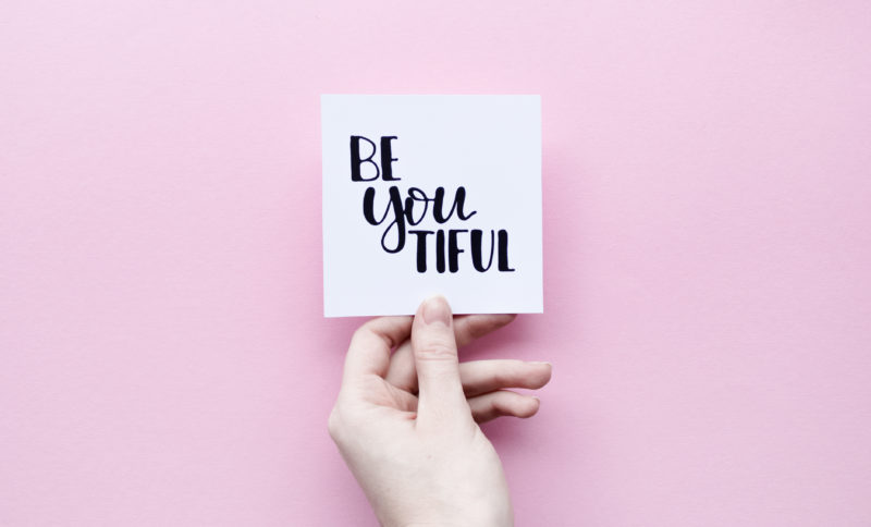 Minimal composition on a pink pastel background with girl's hand holding card with quote - be you tiful