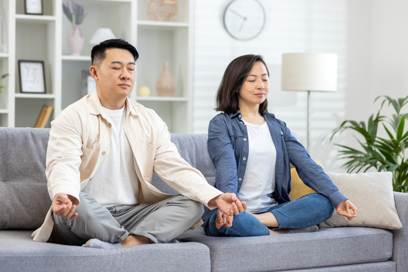 Asian couple at home on a couch meditating; practicing self-care