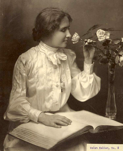 Helen Keller with her hand on a braille book as she smells a rose