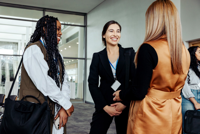 Three businesswomen networking, smiling, and conversing in a professional setting. 