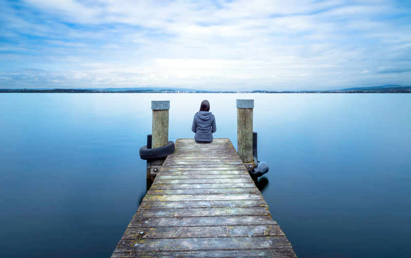 A person sitting at the end of a weathered wooden pier, overlooking a calm, expansive lake. The sky is partly cloudy, and the serene water stretches out towards distant hills, creating a peaceful and reflective atmosphere.