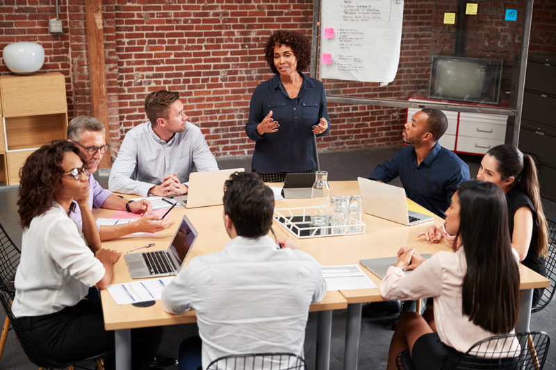 Group of diverse professionals having a meeting in a modern office with brick walls, led by a woman standing and speaking, while others sit around a table with laptops and documents