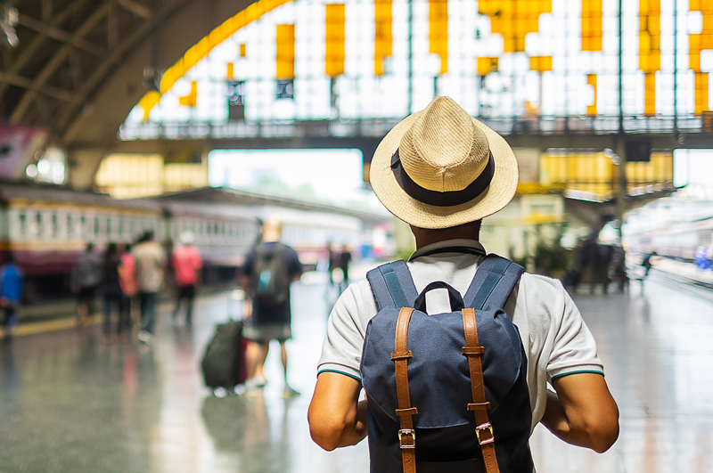 Young man traveler with backpack waiting for train, Asian backpacker with hat standing on railway platform at Bangkok train station. 