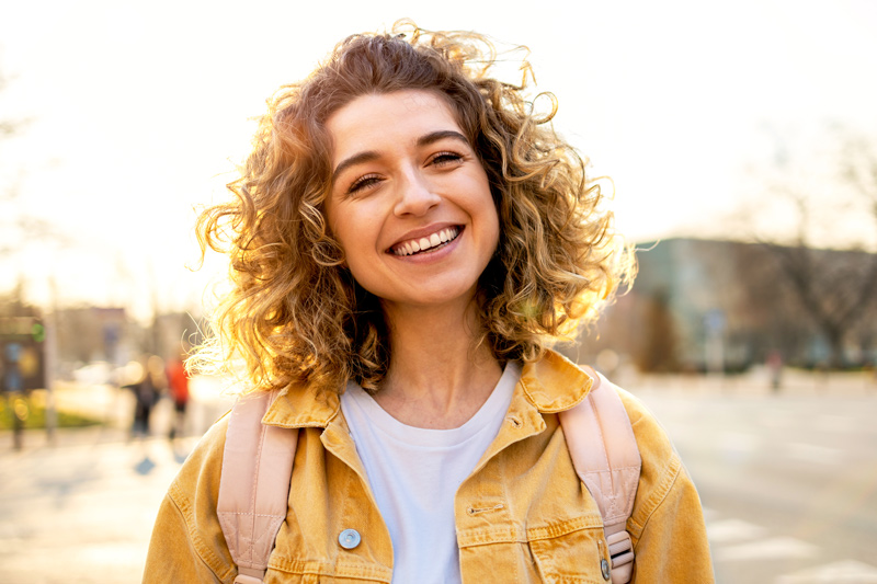 Smiling young woman with curly blonde hair wearing a yellow jacket and a backpack, standing outdoors on a sunny day. Photo for Building Self-Advocacy Skills in Autistic Young Adults