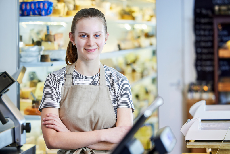 A young woman wearing an apron stands confidently with her arms crossed in a shop, likely a deli or cheese store. She smiles warmly at the camera, with shelves of various products visible in the blurred background, creating a welcoming atmosphere