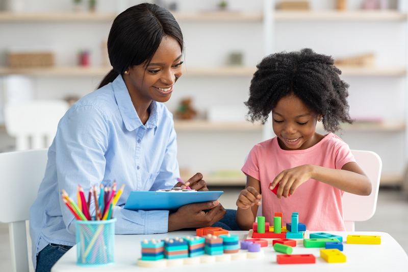 Female psychotherapist friendly pretty young black woman working with little kid at office, sitting by preschooler girl playing with bricks, taking notes and smiling. For Demystifying the Evaluation Process for Your Infant, Toddler, and Preschool Child with Developmental Concerns on Disabilities Education Act