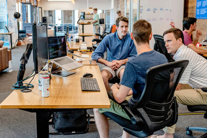 Three men collaborating in an open office, with a laptop displaying code on the desk nearby