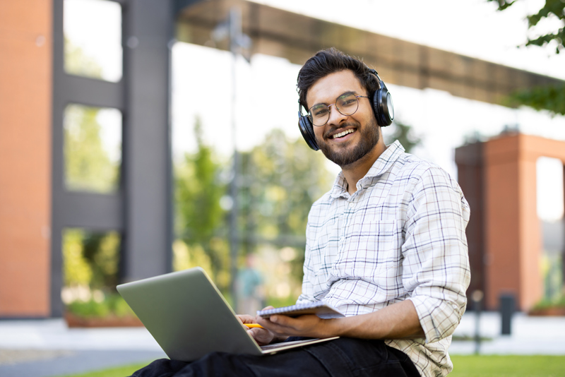 Portrait of an Indian young man wearing headphones sitting on a bench outside, holding a laptop and a notepad on his lap, smiling and looking at the camera