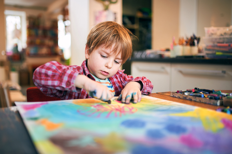 Young boy in a plaid shirt drawing with colorful pastels on a large sheet of paper at home. Focused on creative activity in a cozy art space.