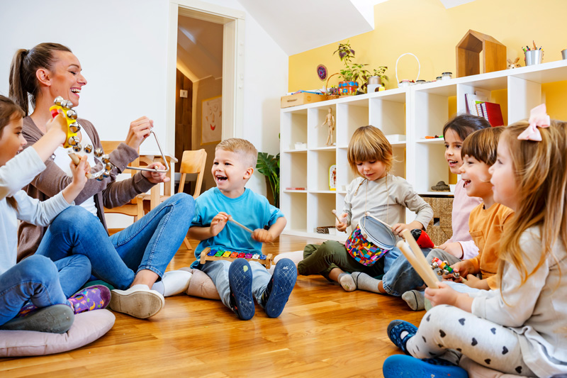 Kindergarten teacher with children sitting on the floor having music class, using various instruments and percussion. Early music education.