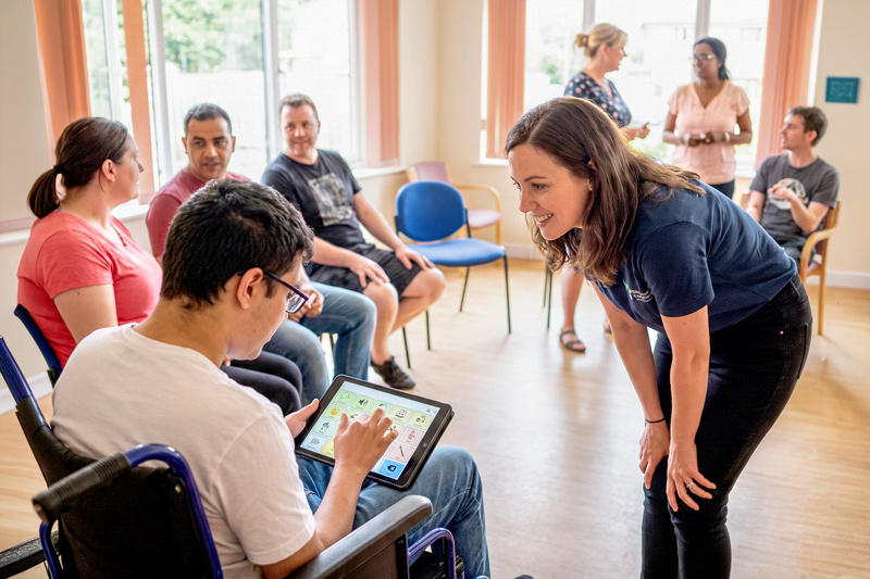 An Ally Speaking With a Man Using an AAC Tablet in a Group Setting